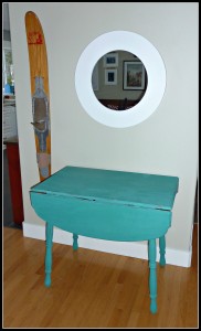 A turquoise little table and a round mirror above it.