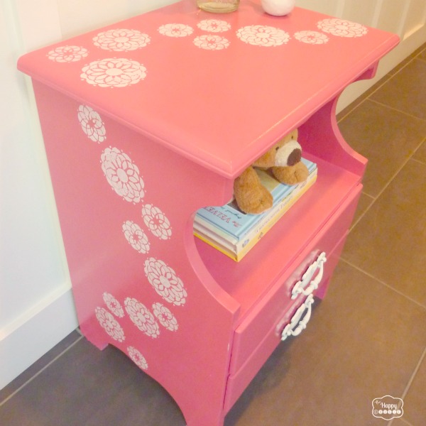 A coral pink nightstand with white flowers and a teddy bear and books in it.