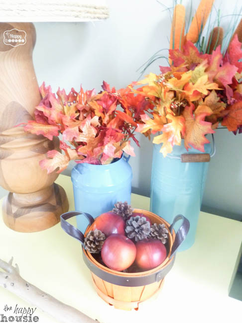Two blue vases with leaves in them, plus a wooden basket filled with apples on the table.