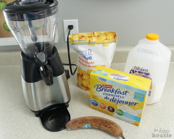 A blender beside milk, a banana, frozen mango and Carnation breakfast essentials.