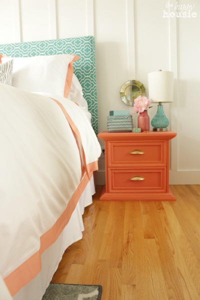 A peach side table beside the bed has lamps and a flower in a vase on it.
