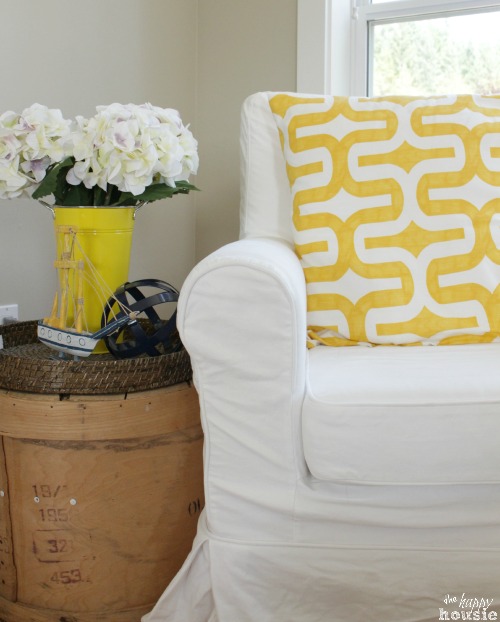A white armchair with a yellow pillow and white flowers beside it.