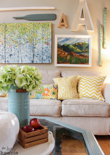 A neutral couch with yellow pillows on it and paintings above it.