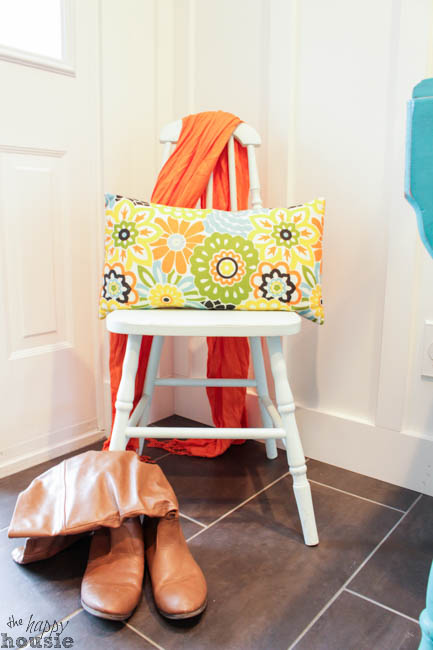 A bright yellow floral pillow on a white chair with boots in front of it, in the foyer.