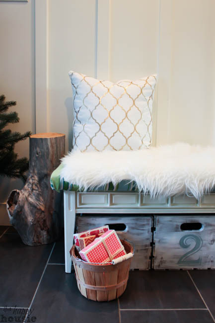 A gold and white pillow on a bench with a basket filled with Christmas presents.