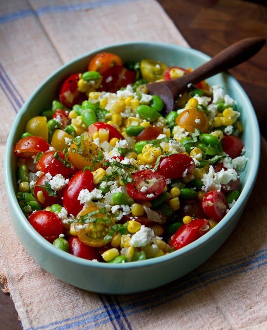 A light green bowl with a wooden spoon in a tomato, corn, feta salad.