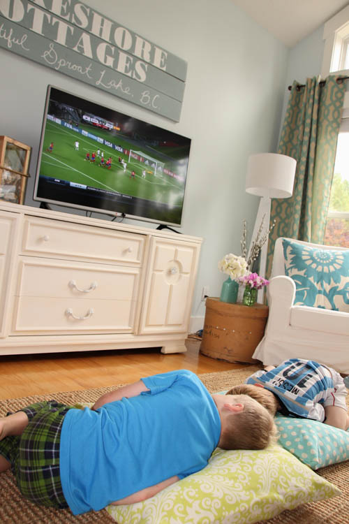 Two boys on the pillows on the floor with sports on the TV.