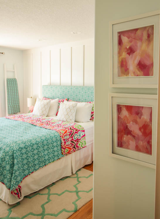 A queen sized bed with a white wall behind it, a patterned bedspread in turquoise and shades of pink.