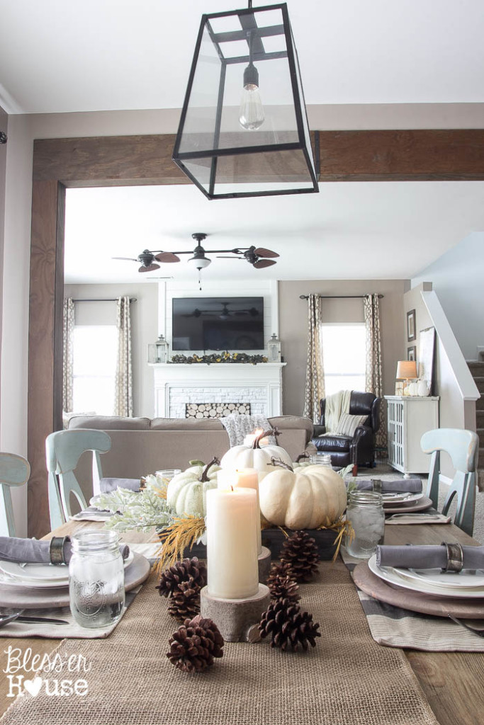 There is a neutral runner on the table with candles, pine cones, and white pumpkins.