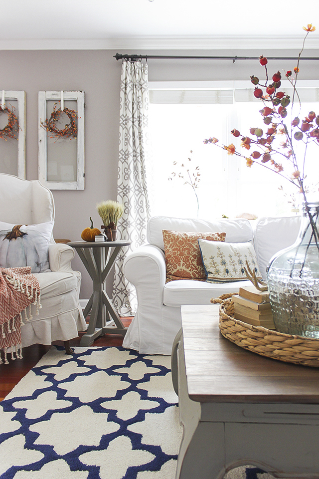 A blue and white rug is on the floor with a white couch and armchair.   Plus a clear jar with red berries on the table.