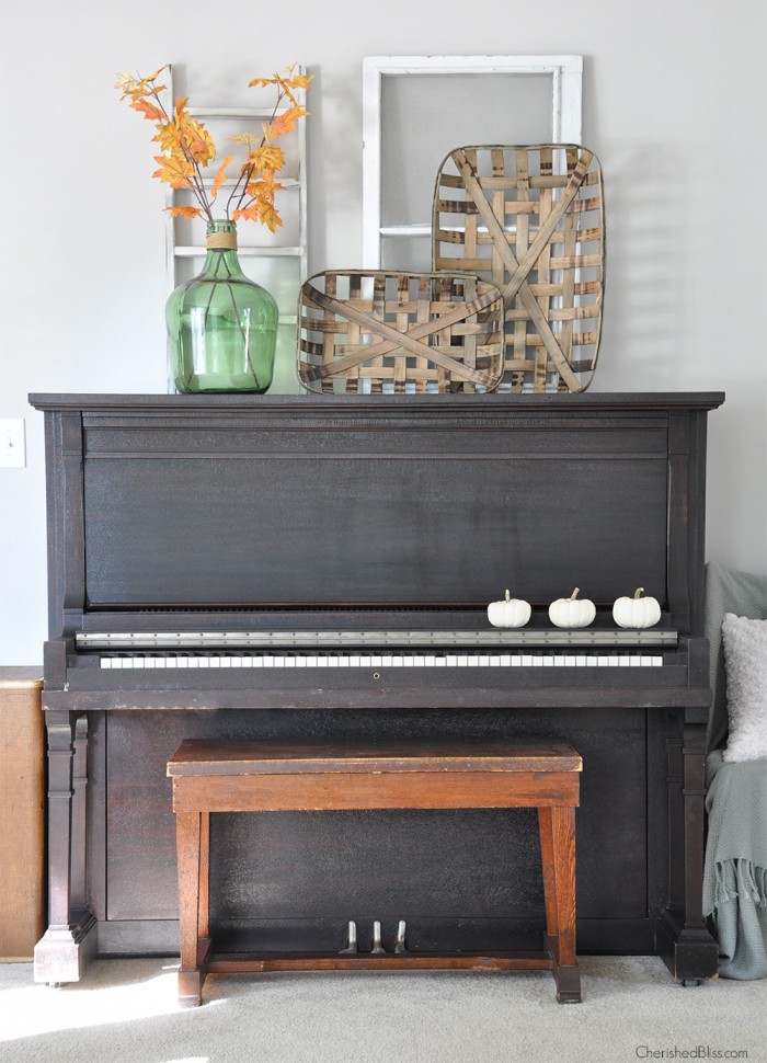 An old piano has three mini white pumpkins on it and on top of the piano is a green glass jar with yellow leaves.