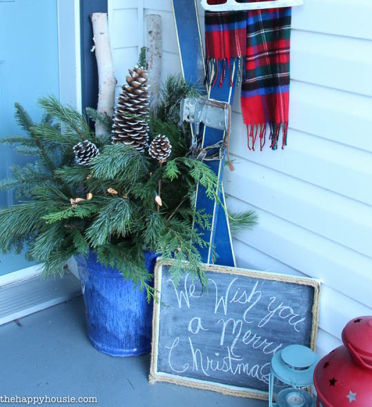 The greenery display in the corner of the porch beside skis and a sign that says we wish you a Merry Christmas.