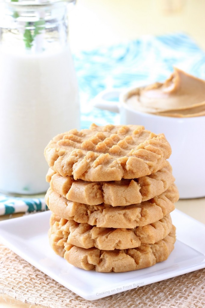 A stack of peanut butter cookies on a plate on the table beside a cup filled with peanut butter.