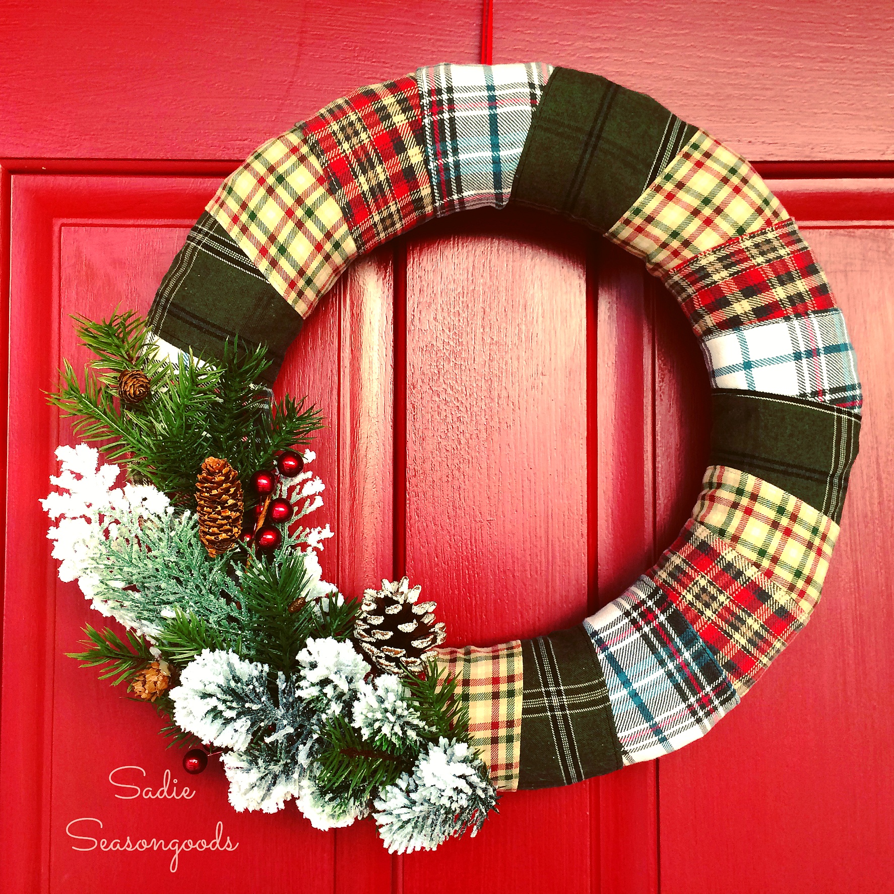 A red door with a flannel sheet wrapped around a wreath and pinecones and holly and berries.