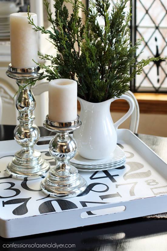 A tray with black numbers on it and candlesticks and a white vase filled with greenery.