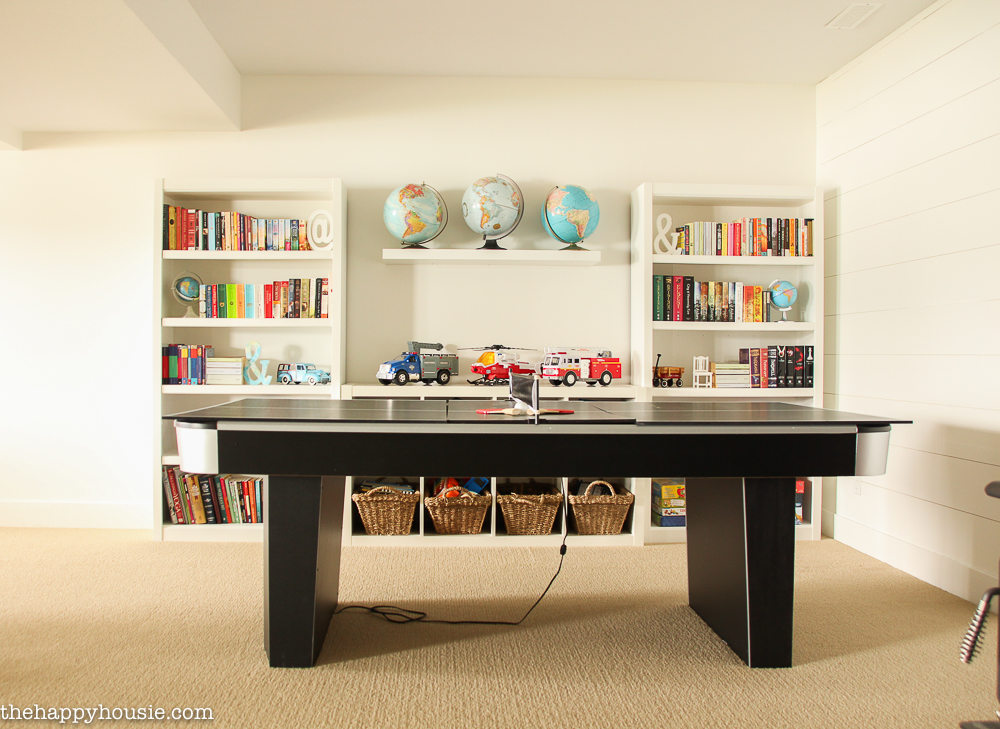 A ping pong table is beside a bookshelf in the basement.
