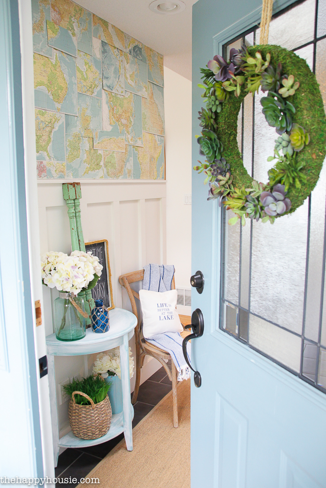 A light blue door with a green and succulent wreath on it, the door is ajar showing the inside hall.