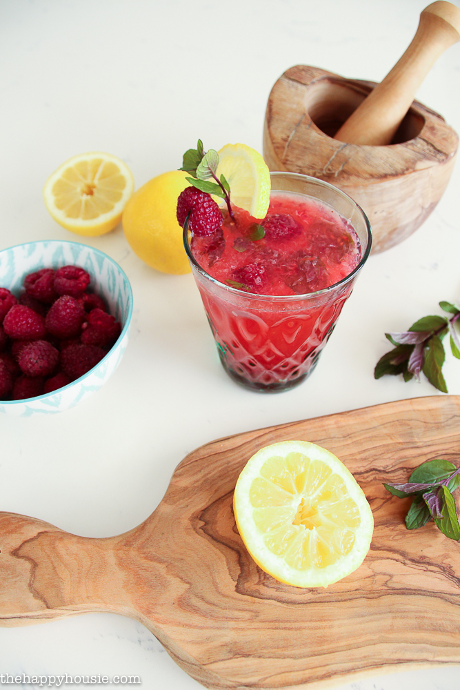The raspberry mint lemonade in a clear glass with the lemons, raspberries beside it.
