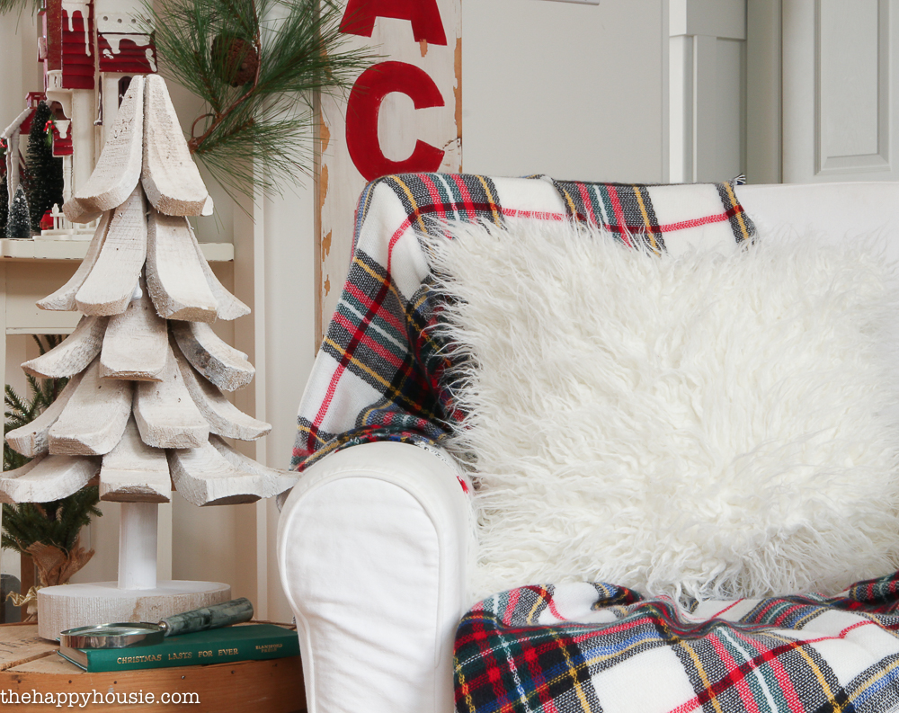 A white fur pillow on a white armchair with a plaid throw blanket.
