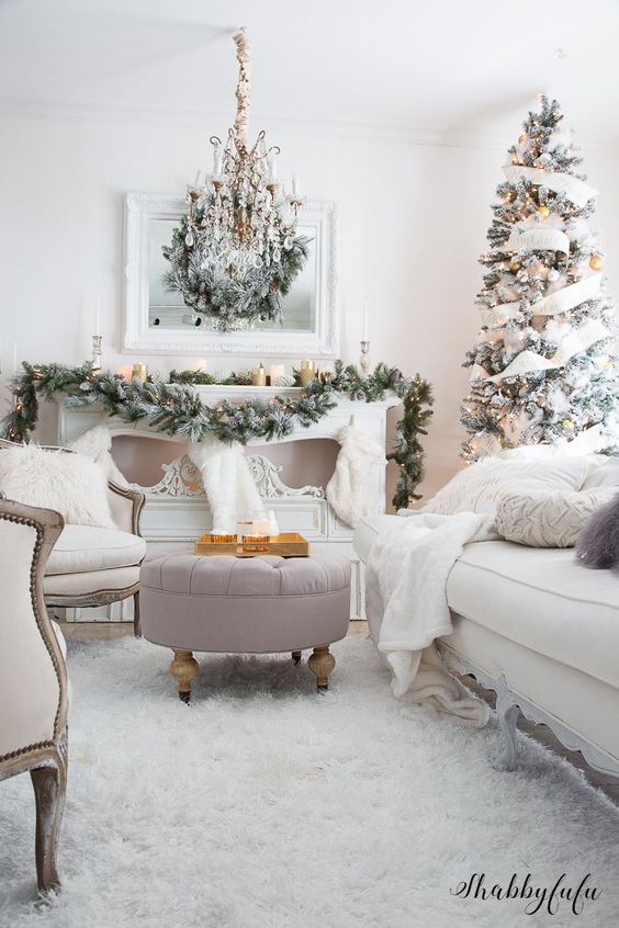 A large chandelier in a white living room with a blush ottoman and a White Christmas tree in the corner of the room.