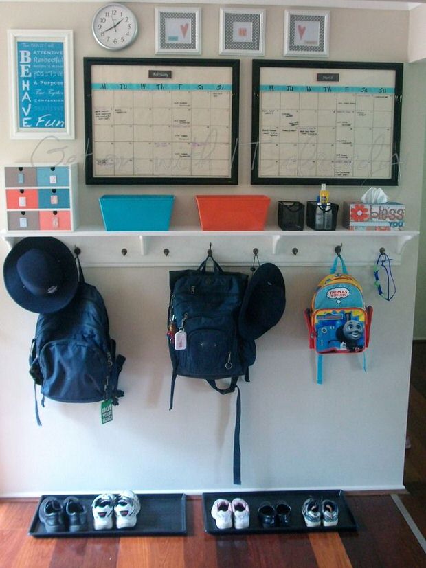 A white wall with backpacks on hooks and schedules on the wall, beside a clock.