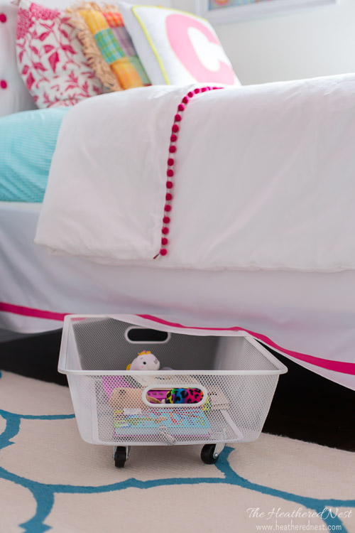 A white bedspread with pink details an a small rolling under the bed cart.