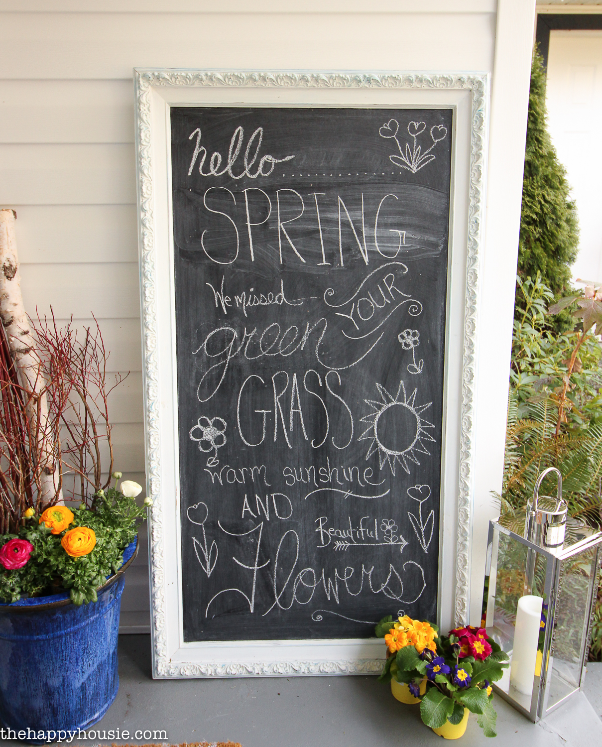 There are pots of flowers by the chalkboard on the porch.