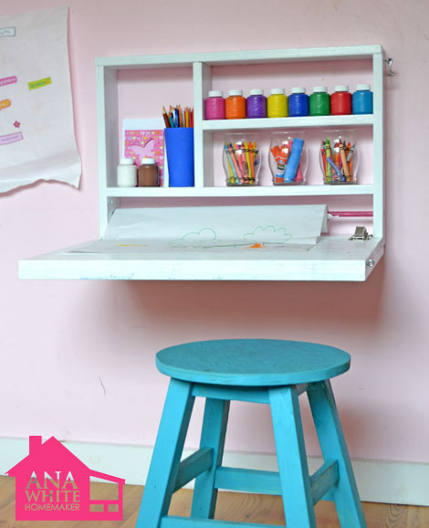 A small pull down white shelf with paints and crayons in it.