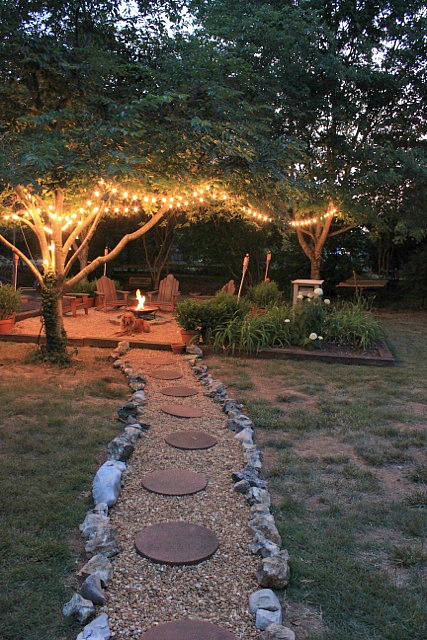 A path in a backyard leading to a fire pit with trees around it and lights on the trees.