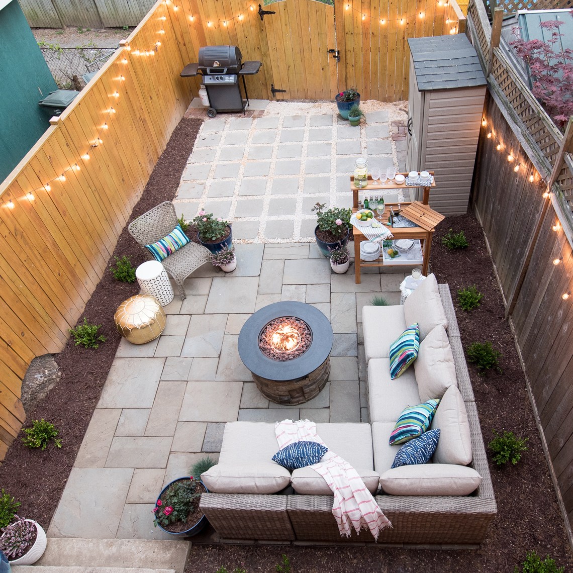 An aerial view of a backyard porch with a fence around the yard and lights on the fence and a round lit fire pit.