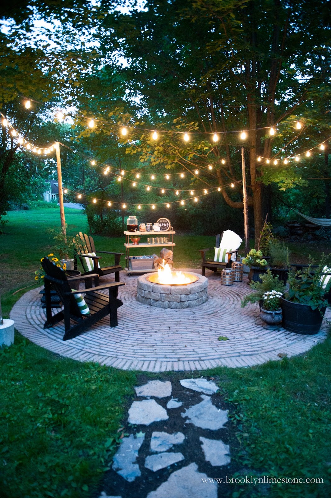An outdoor round flat to the ground rock work patio with chairs around the fire pit and a string of light above it.