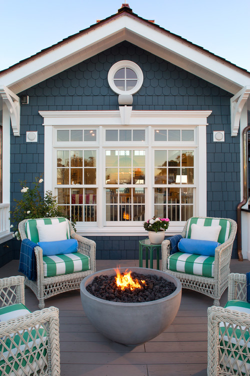 A blue and White House with a fire pit on the deck and green and white cushioned chairs are around it.