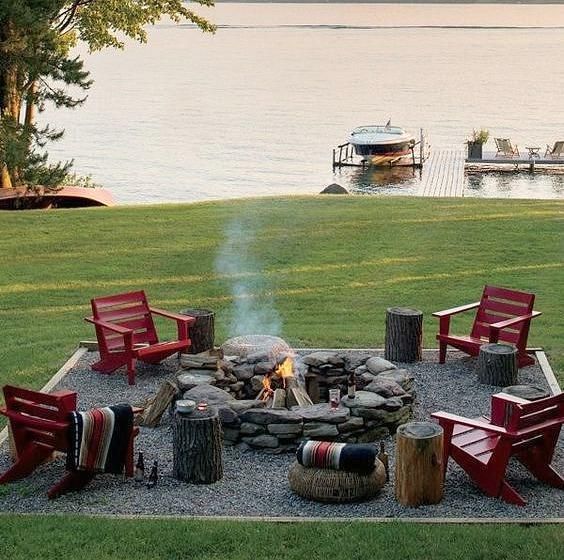 A lake with a boat dock and a fire pit on the lawn with red chairs.