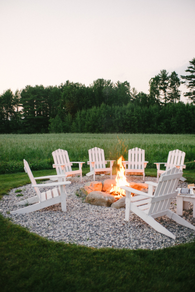A large grassy field with a circle of rocks in the middle of it and fire pit on the rocks with white chairs surrounding the fire pit.