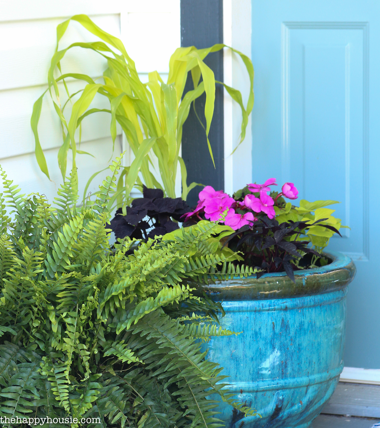 There are potted pink plants and green plants in a blue planter on the front porch.