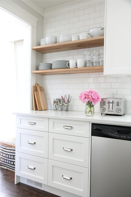 A white kitchen with pink flowers on the counter and shelves with dishes above the counter.