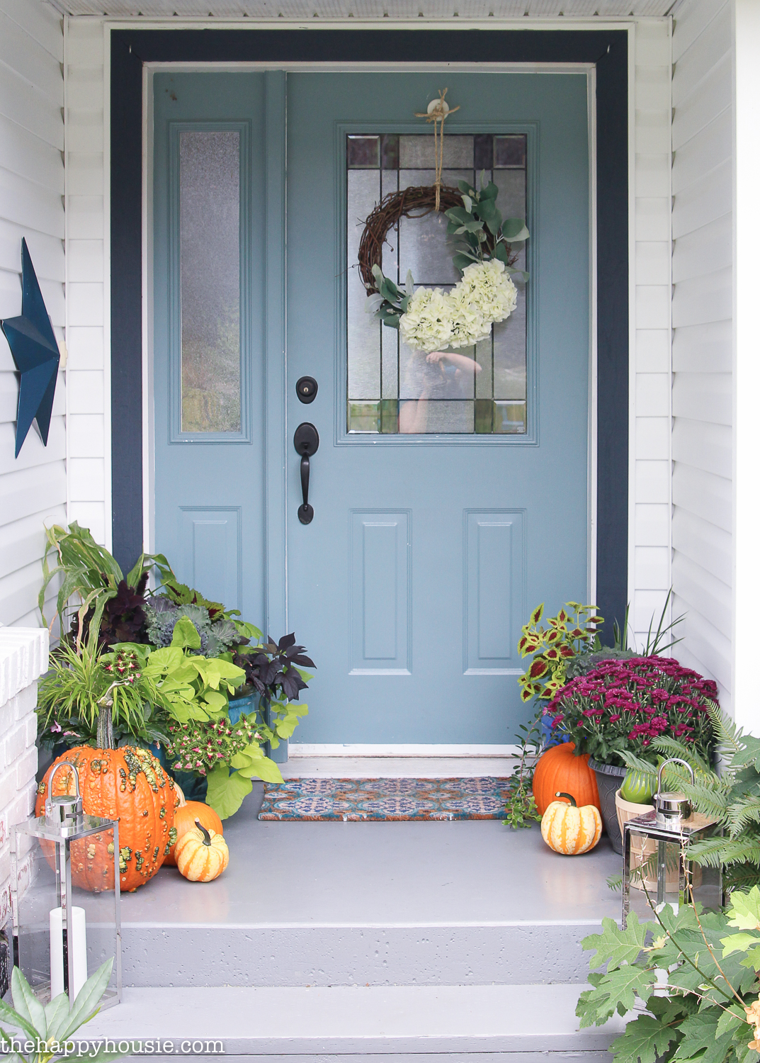 A light blue front door surrounded by flowers, greenery and pumpkins.   There is a fall wreath on the door.