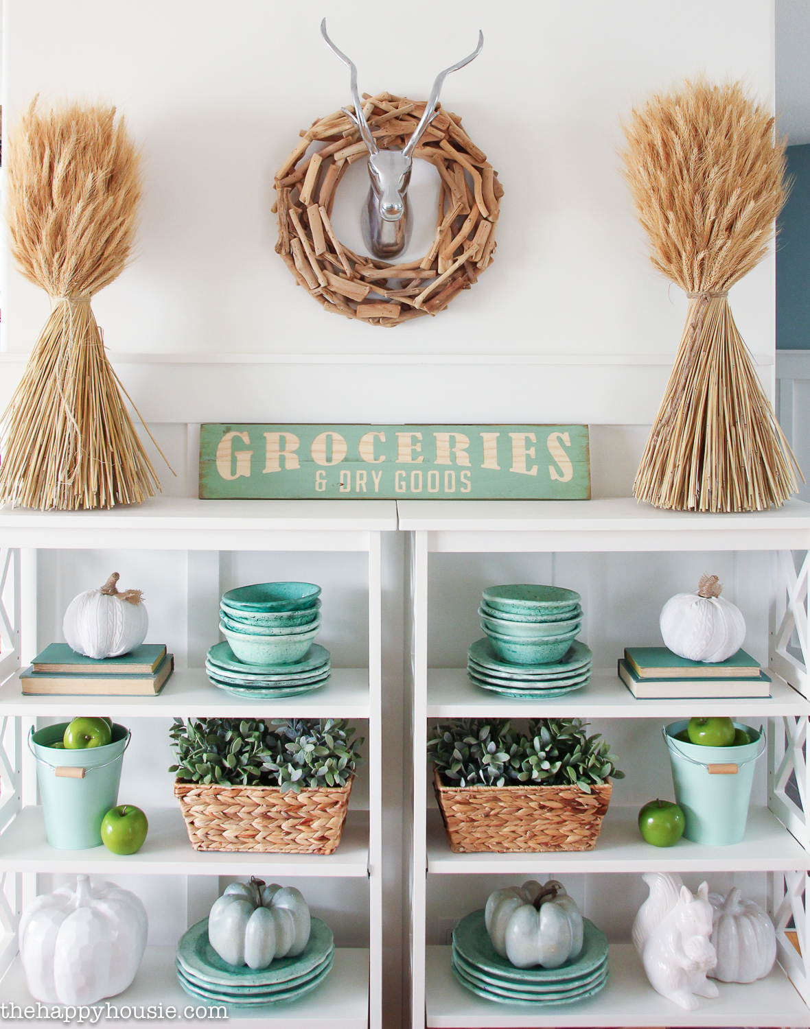 A white shelving unit is in the dining room with stalks of wheat and a wreath on the wall with antlers.