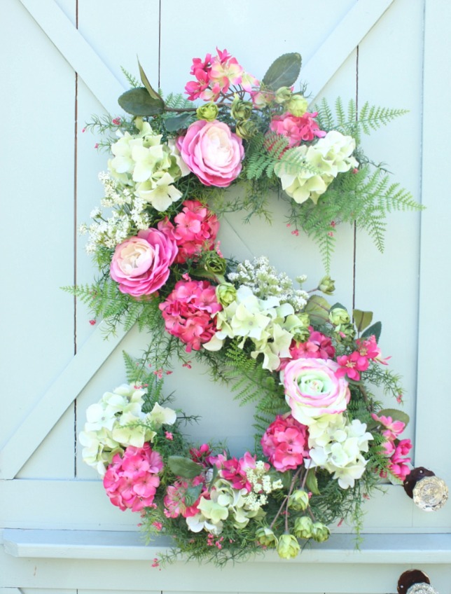 A large floral wreath forming an S in white, pink flowers with greenery, hanging on a barn door.