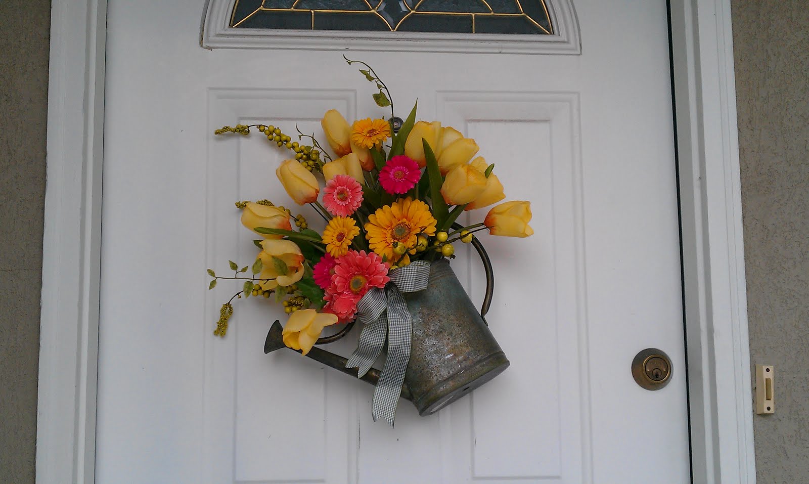 A watering can with yellow and pink flowers in it hanging on a white door.