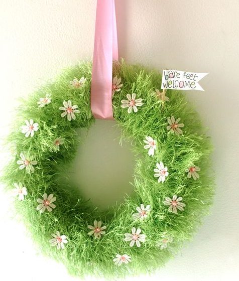 A fluffy green wreath with small pink and white flowers on it. There is a pink ribbon around it hanging on a wall.