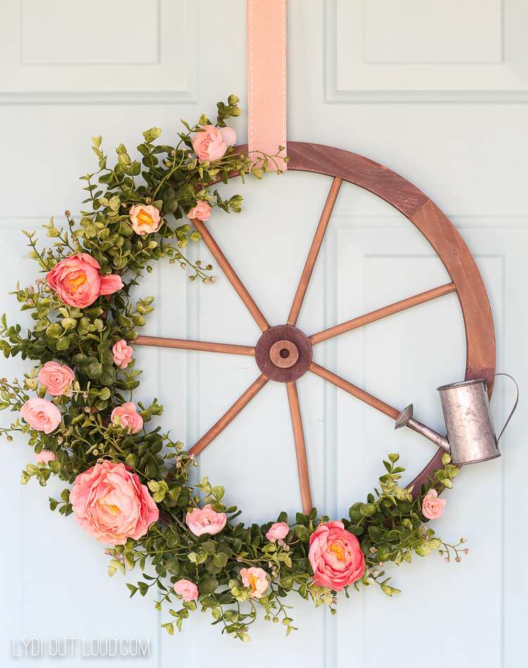 A wooden circle with wooden spokes, a watering can and flowers with green vines on the summer wreath.