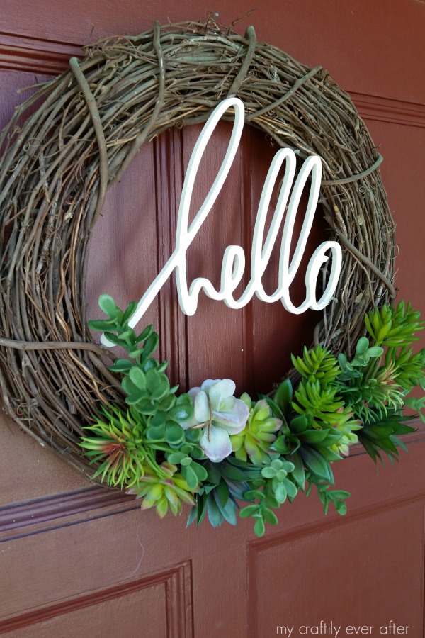 A grapevine wreath with greenery on the bottom and the word hello in white on the wreath.