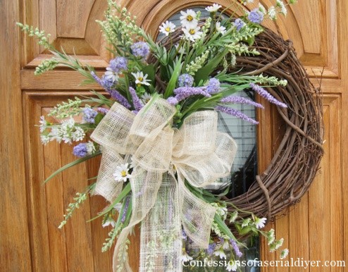 A wooden door with a twig wreath and lavender, daisy flowers. Plus a large white bow on the wreath.