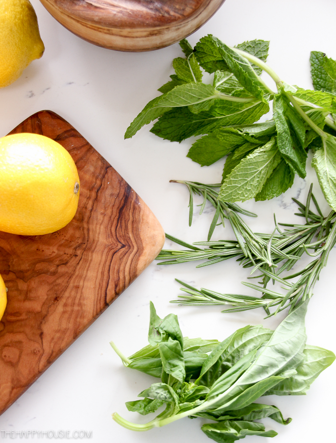 Herbs on the counter beside a wooden cutting board with a lemon on it.