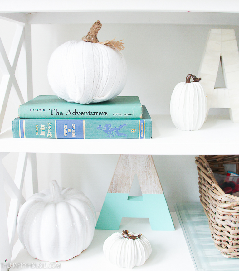 The white sweater pumpkins on the open shelf sitting on books and beside baskets.
