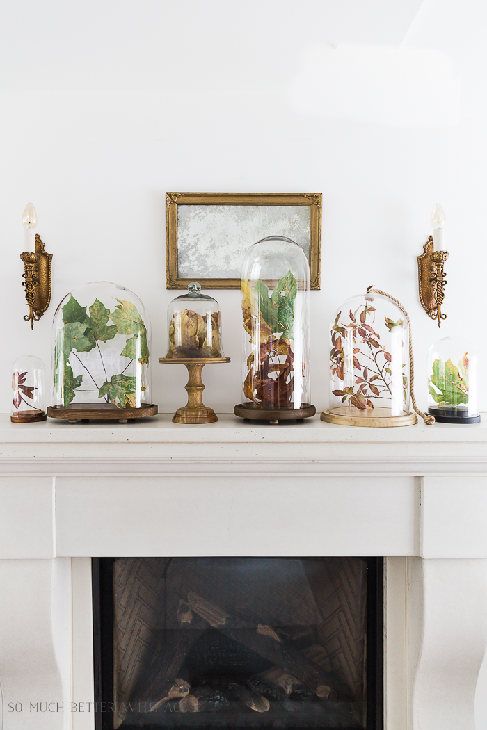 A white fireplace mantel with cloches filled with autumn leaves.
