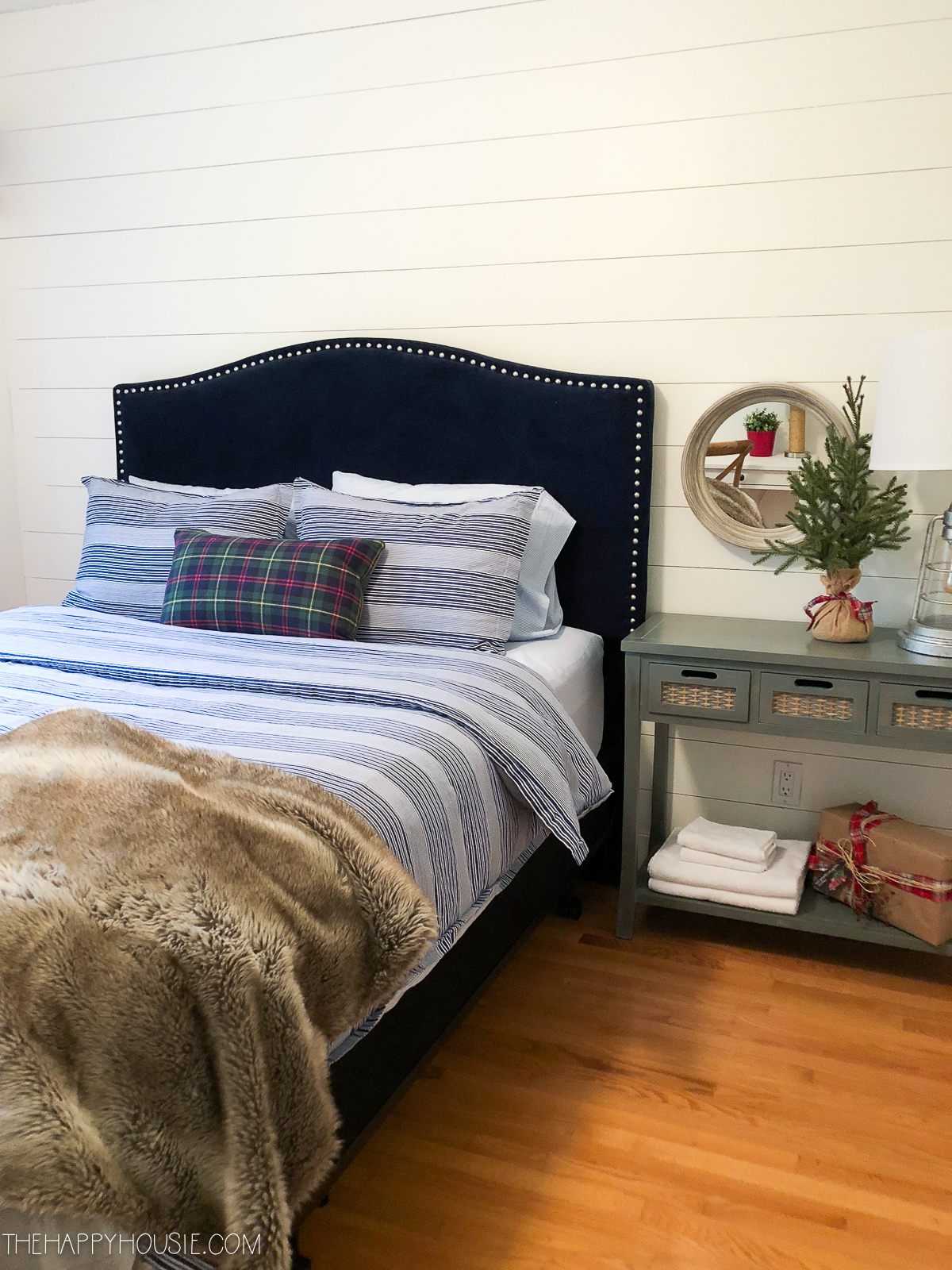 A fabric headboard with rivets on it in a dark blue, with a small table beside it and a small Christmas tree on it.