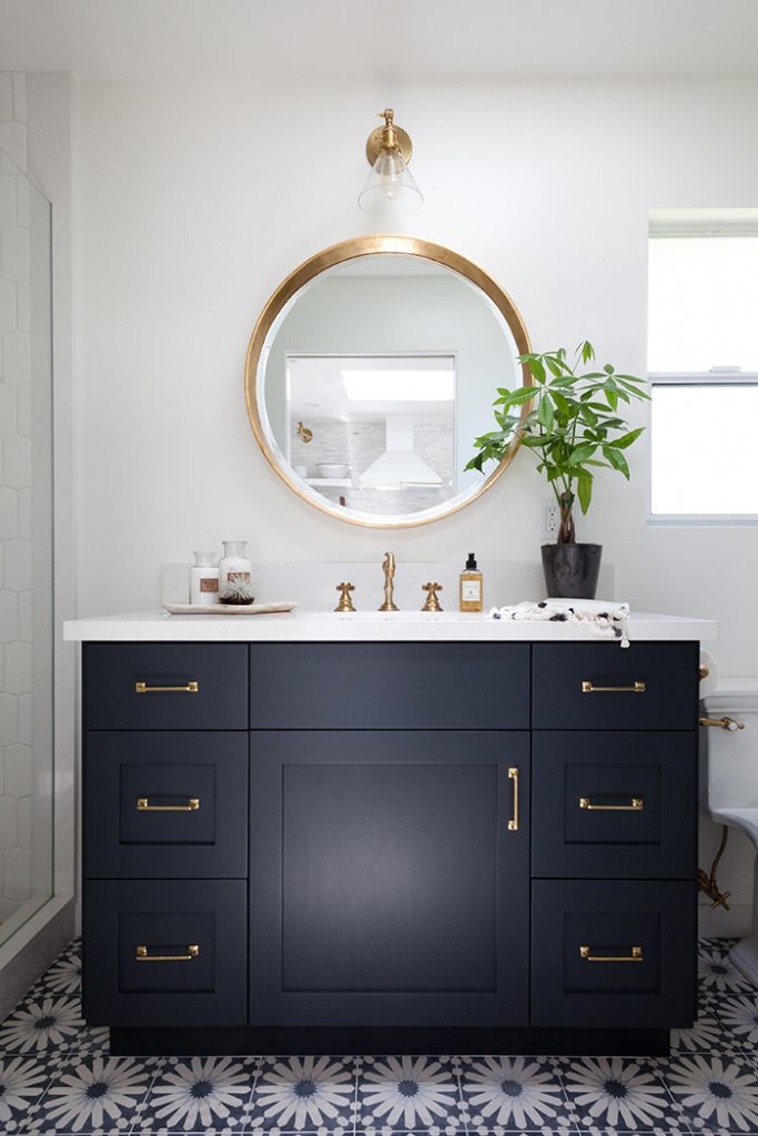 A round brass mirror in the bathroom above the dark blue vanity.