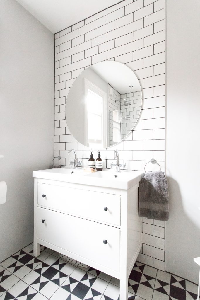 An all white bathroom vanity with a round mirror and diamond geometric black and white floor.